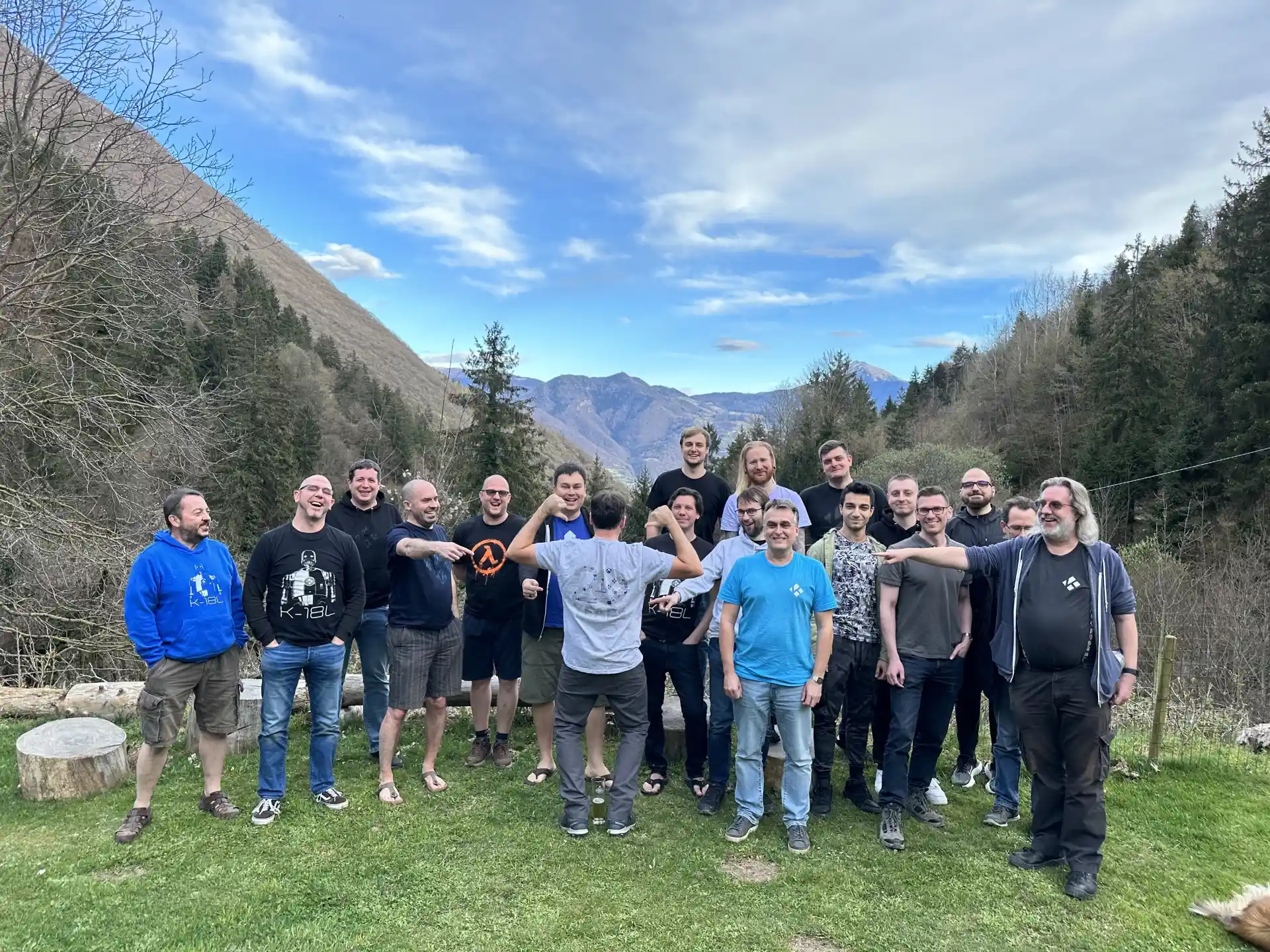 Team Kodi, 2023 - Kodi team members at Devcon 2023. in northern Italy. A group of people stand, spread out, facing the camera. Grass in front, but mountains soar in the background behind this motley crew of technological reprobates. One team member has his back turned to the camera, while people point to the logo on the back of his shirt: the numbers 20, signifying the 20th anniversary of XBMC/Kodi, but made up of the names of all the people and companies that have helped us make this awesome software happen.