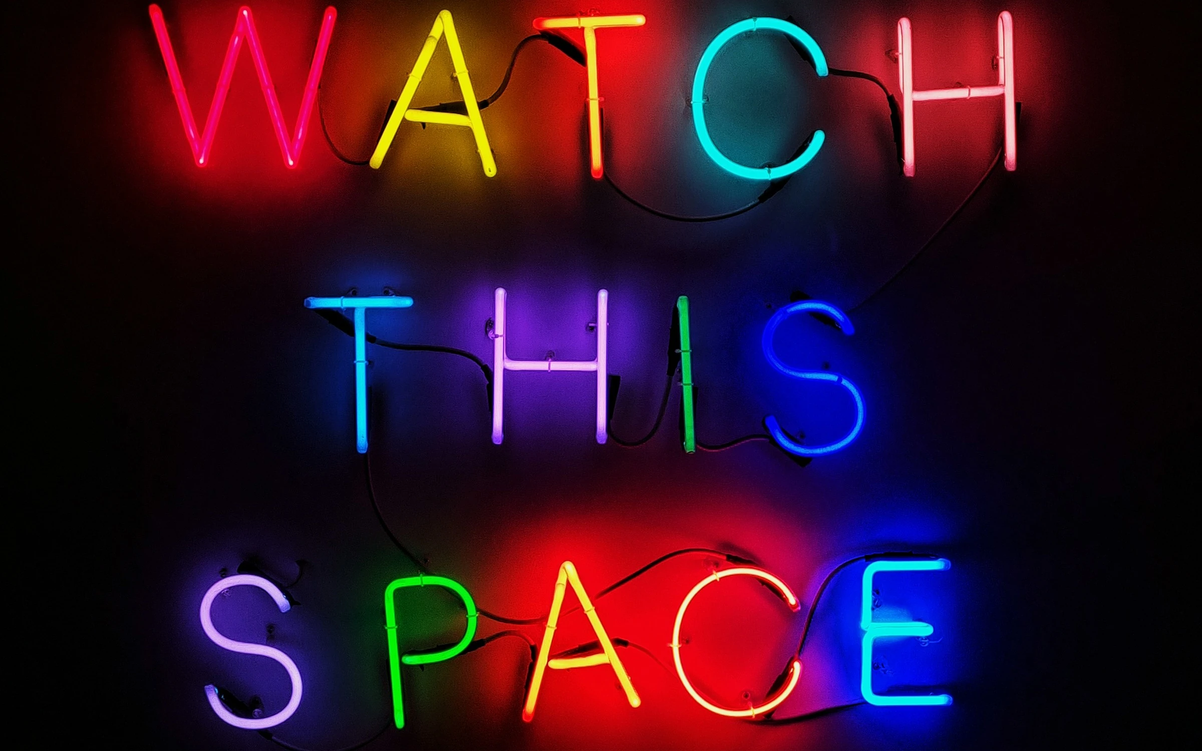 A glowing neon sign that says "Watch This Space", with each letter coloured differently.
